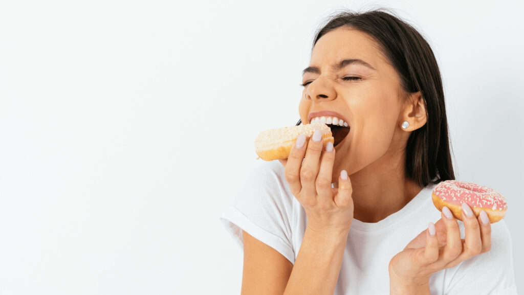 Mujer morena muerde con fruición un donut con glaseado beige y espolvoreado