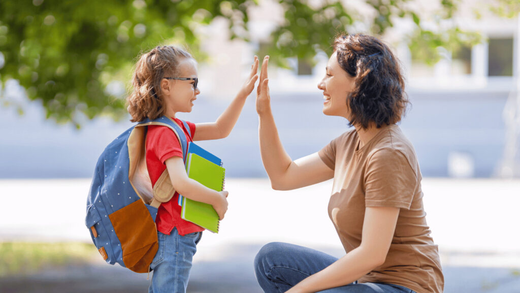 Une mère brune et un petit écolier avec des lunettes, des livres d'école et un sac à dos bleu se font un highfive en souriant