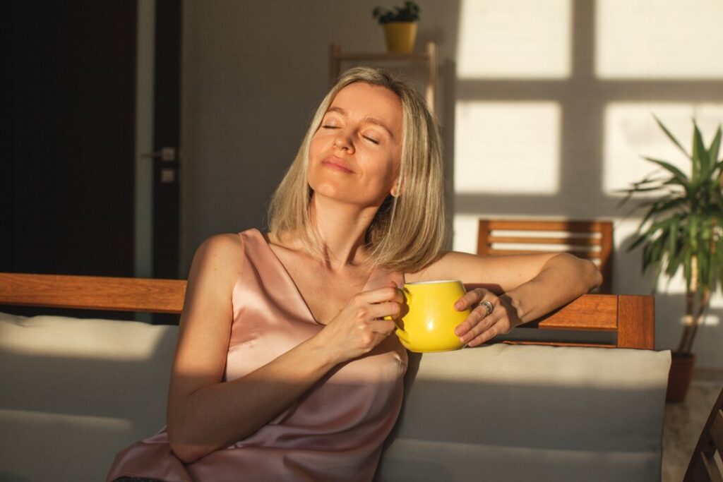 Une femme est assise sur un canapé, le soleil brille sur son visage. Elle tient une tasse à la main