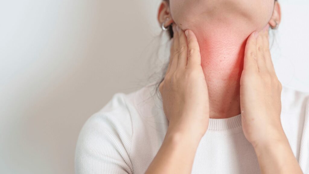 woman with white t-shirt, dark braid on white background holds both hands to her thyroid gland which is marked red, she has hashimoto's disease