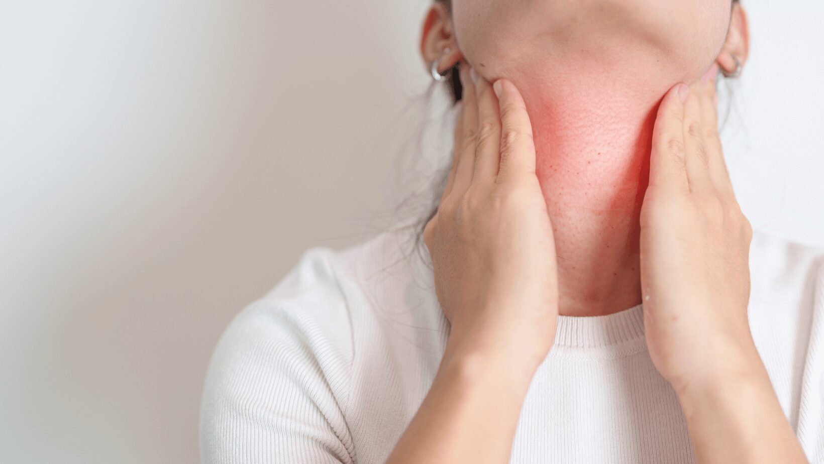 une femme avec un t-shirt blanc, une tresse foncée sur un fond blanc se tient les deux mains sur sa thyroïde qui est marquée en rouge, elle a l'hashimoto