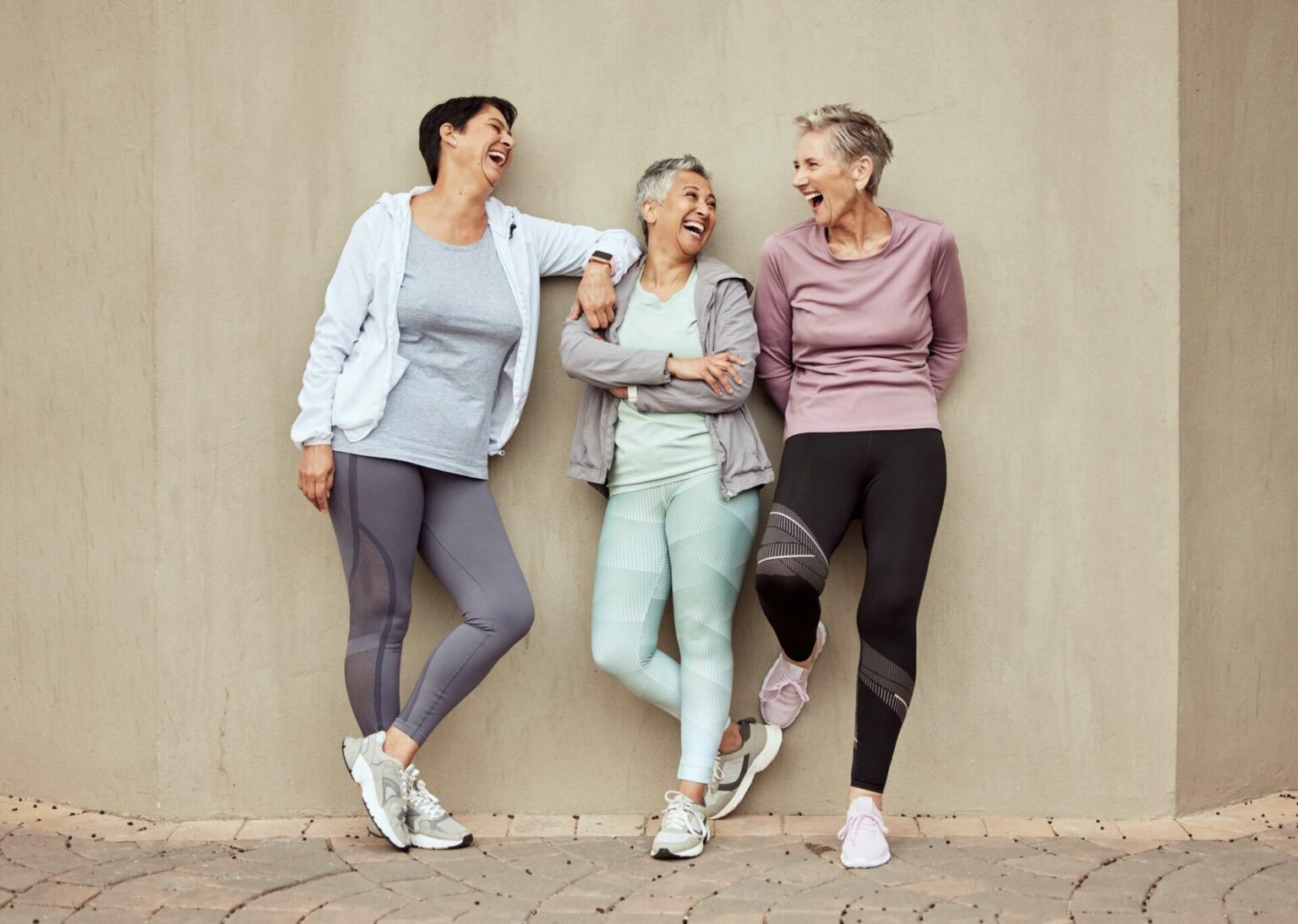 three middle-aged women with short hairstyles stand together in front of a beige wall and laugh at each other